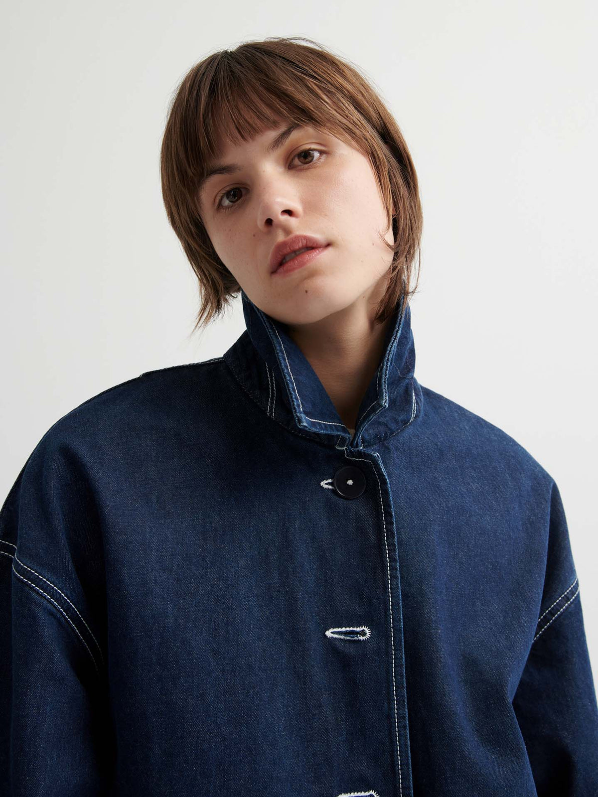 Person with short brown hair wears the Portrait Jacket – Indigo, a dark blue boxy-fit jacket with white stitching, while looking at the camera against a plain background.