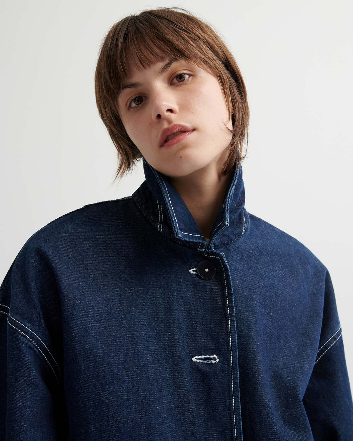 Person with short brown hair wears the Portrait Jacket – Indigo, a dark blue boxy-fit jacket with white stitching, while looking at the camera against a plain background.