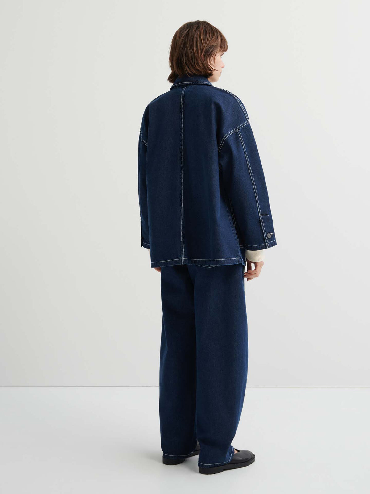 Person standing in a white room, facing away, wearing the Portrait Jacket – Indigo with matching wide-leg jeans and black shoes.
