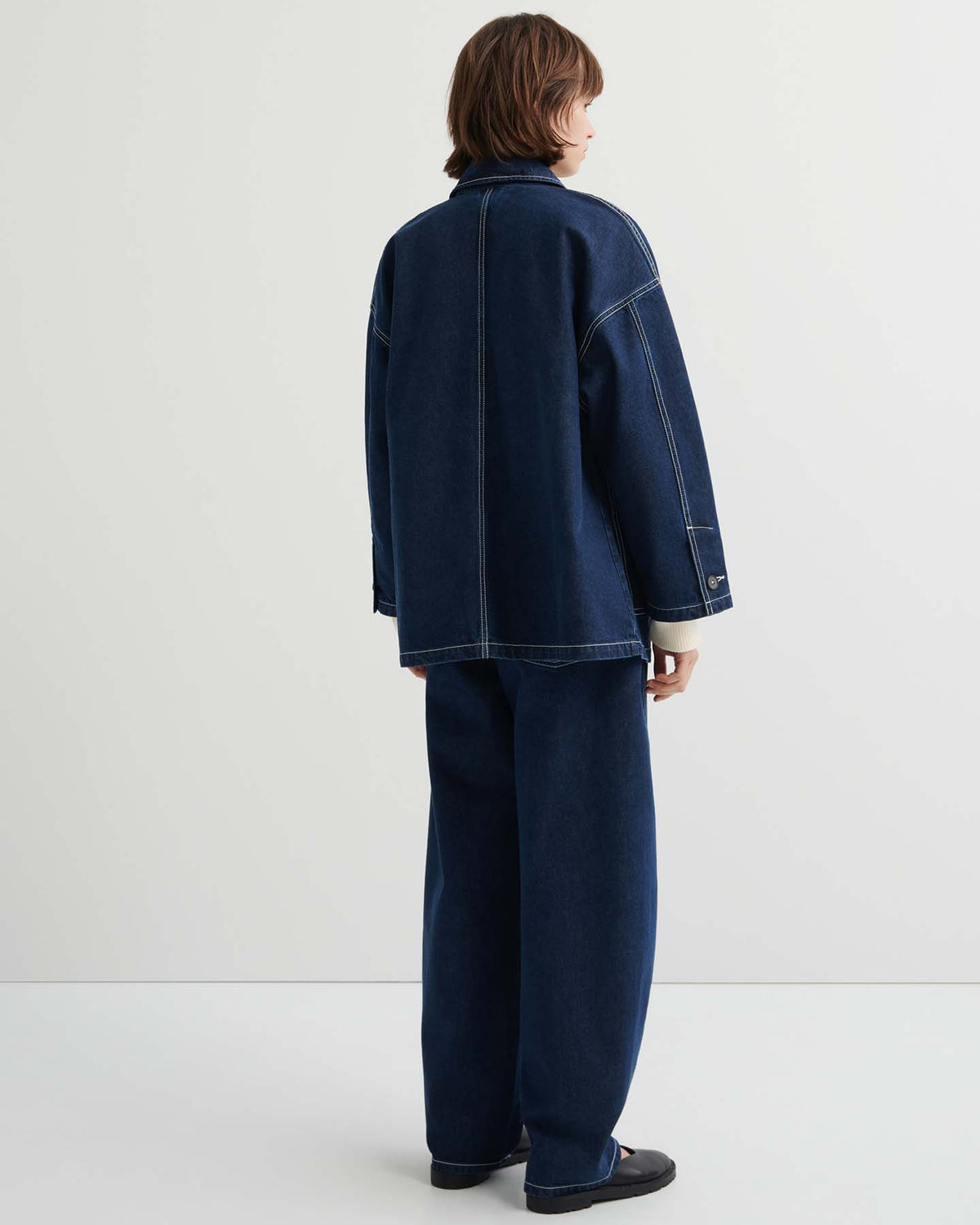 Person standing in a white room, facing away, wearing the Portrait Jacket – Indigo with matching wide-leg jeans and black shoes.