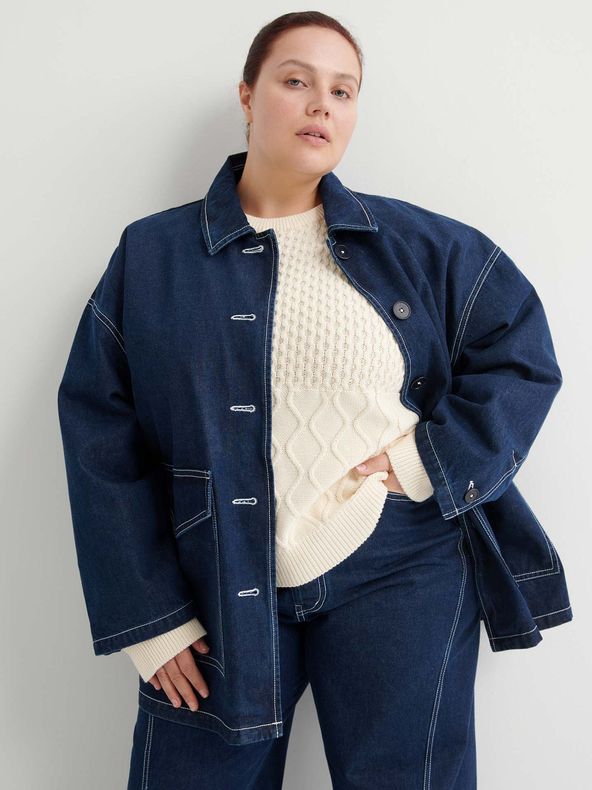 A woman stands against a white wall wearing the Portrait Jacket – Indigo, which has a boxy fit, paired with matching pants and a cream-colored textured sweater.