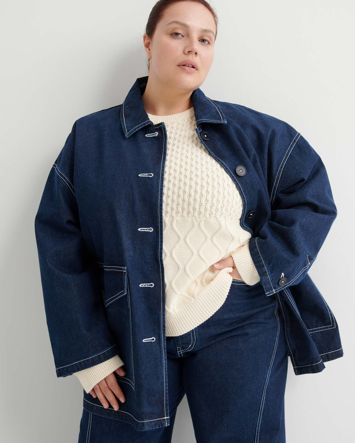 A woman stands against a white wall wearing the Portrait Jacket – Indigo, which has a boxy fit, paired with matching pants and a cream-colored textured sweater.