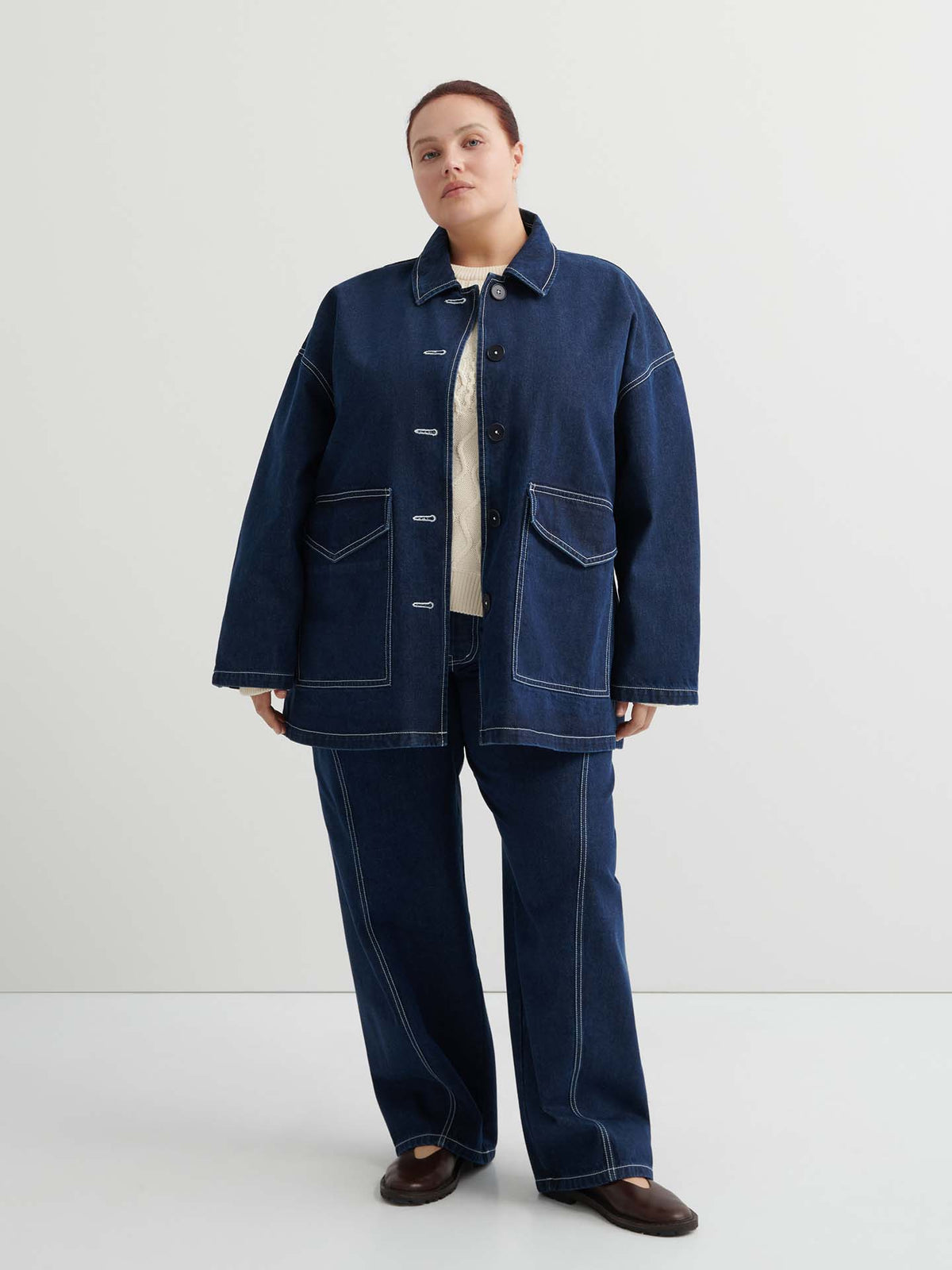 A person stands against a plain background wearing the Portrait Jacket – Indigo, a dark blue boxy fit jacket with white stitching, matching denim pants, and brown shoes.
