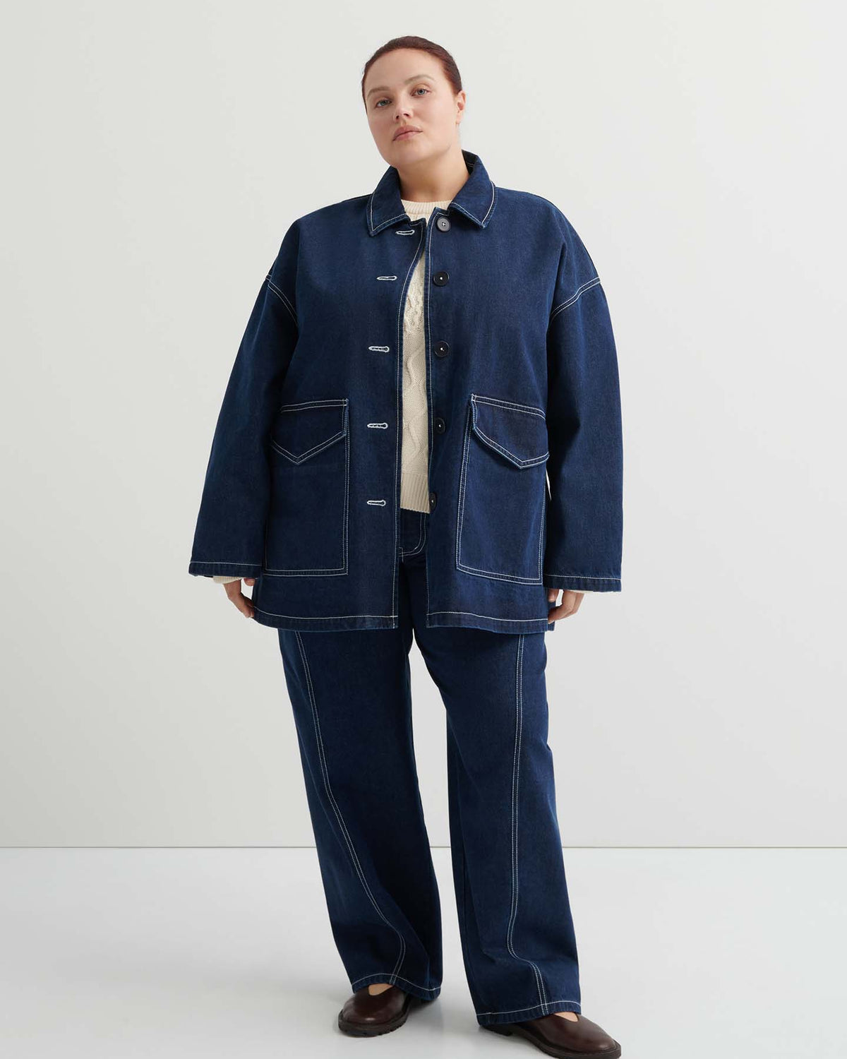 A person stands against a plain background wearing the Portrait Jacket – Indigo, a dark blue boxy fit jacket with white stitching, matching denim pants, and brown shoes.