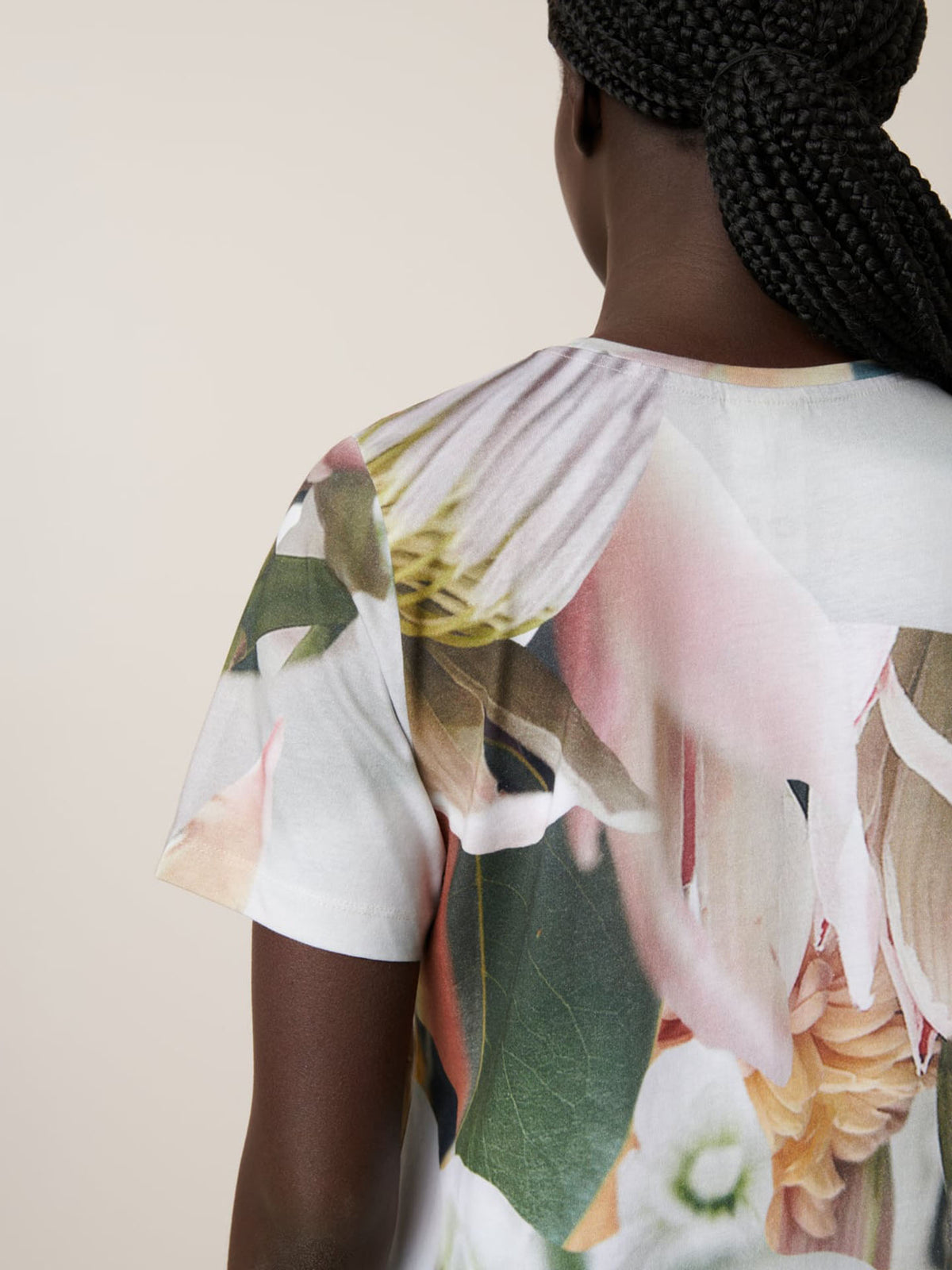 A person with braided hair, seen from behind, wears the Kowtow Protea Tee—a white t-shirt with a large, colorful floral print and an oversized fit. For the perfect style, view the size guide for Protea Tee measurements.