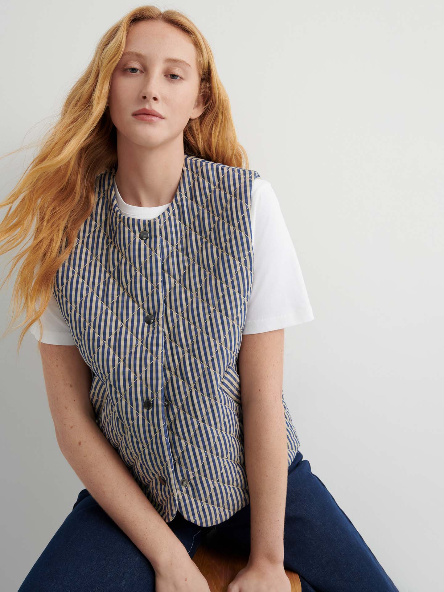 A woman with long red hair layers a white T-shirt under the Quilter Vest – Landscape check, featuring blue-and-tan stripes, as she sits against a plain light background.