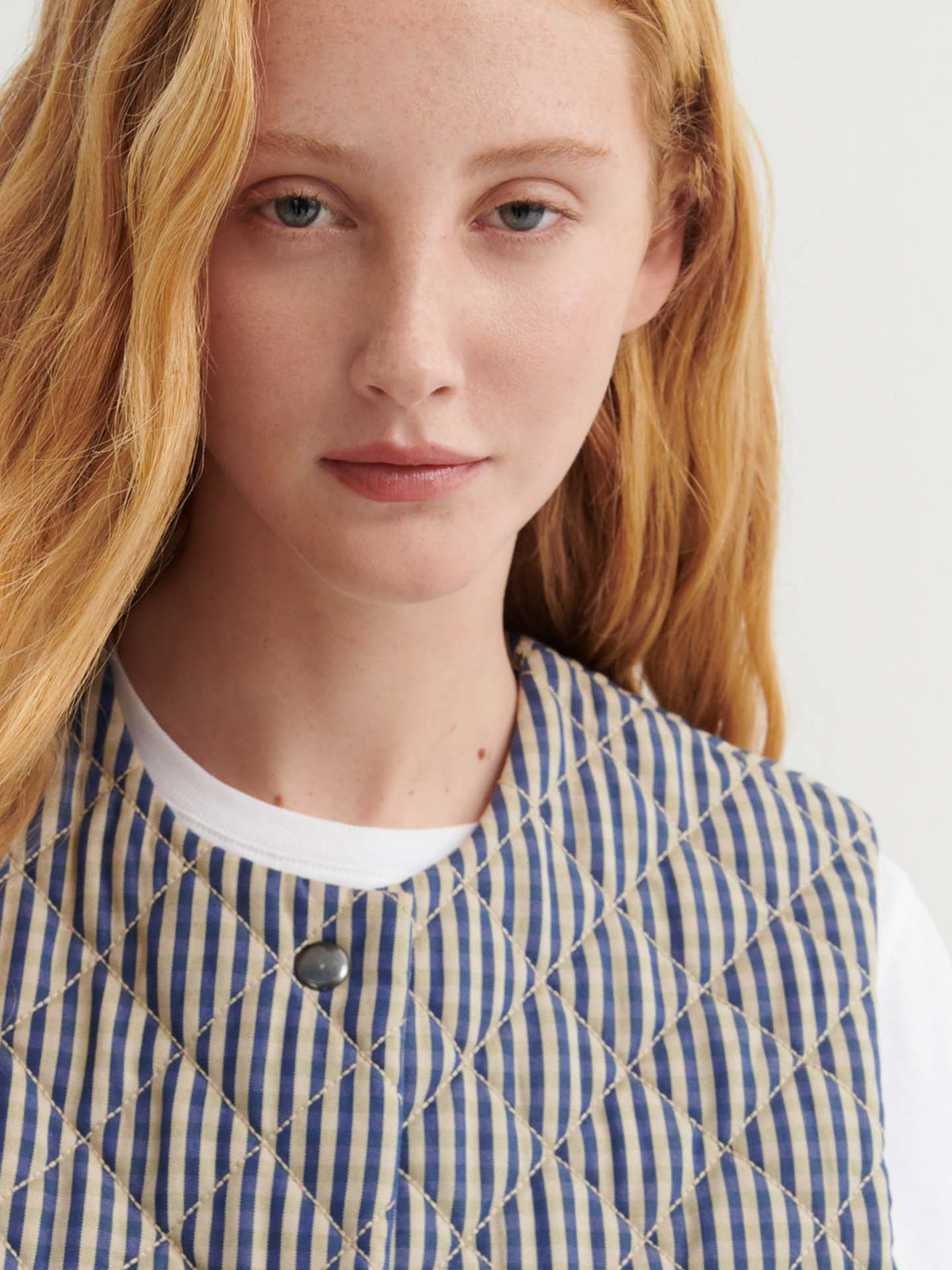 A woman with long reddish-blonde hair wears the Quilter Vest – Landscape check, a blue and beige striped, quilted vest in a relaxed fit over a white shirt, looking directly at the camera.