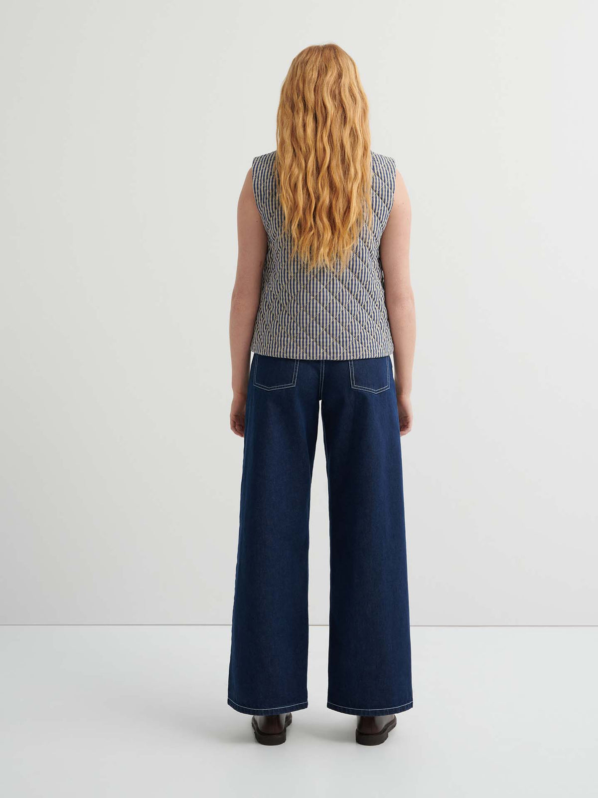 A person with long wavy hair, wearing the Quilter Vest – Landscape check, stands facing away from the camera against a plain background.