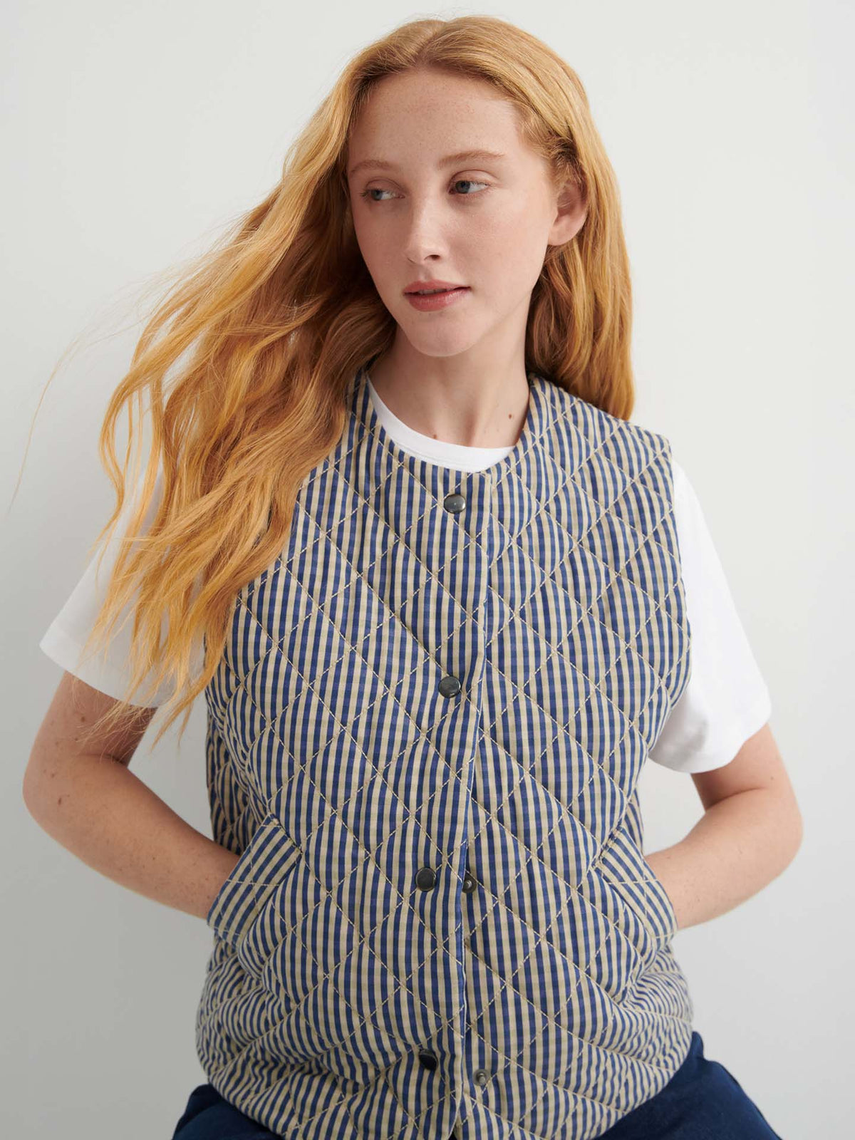 A woman with long red hair wears the Quilter Vest – Landscape check, a blue and beige striped quilted vest in a relaxed fit, over a white t-shirt, looking to her left against a plain background.