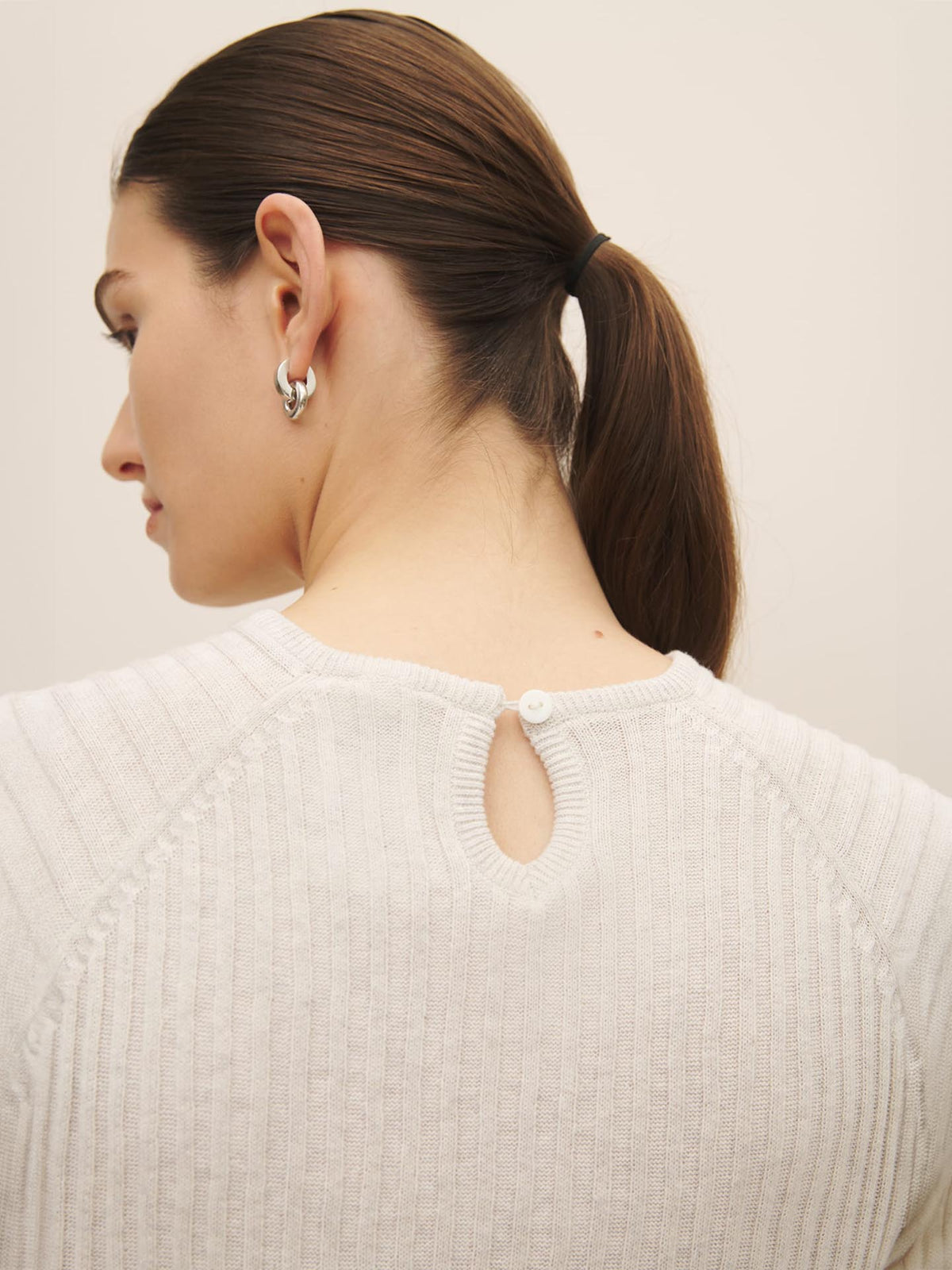 Woman with a ponytail wearing a white sweater, ethically made with Fairtrade organic cotton, featuring a button detail at the nape, (Quinn Top – Light Marle) by Kowtow.