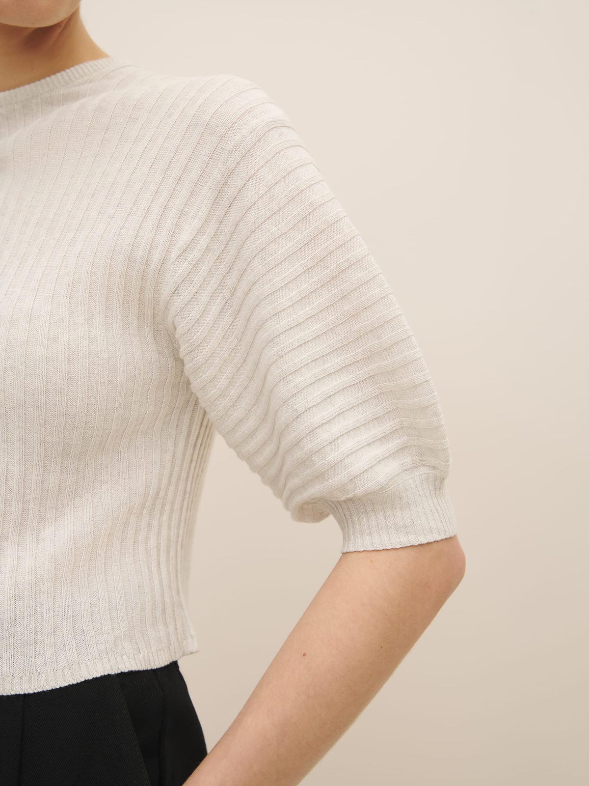 A person wearing a Kowtow Quinn Top - Light Marle, an ethically made white ribbed sweater with a three-quarter sleeve, standing against an off-white background.