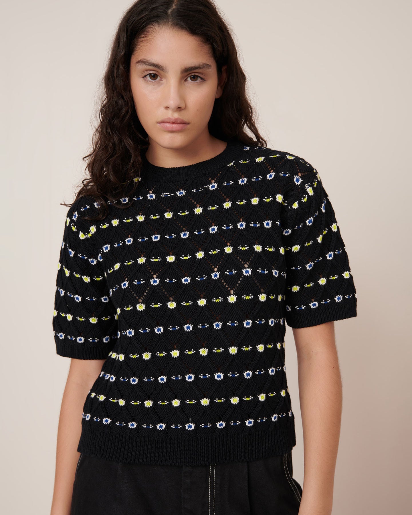 A woman with long dark hair wears the Kowtow Rhode Top – Daisy Jacquard, a black short-sleeve knit sweater with a yellow and white pattern, paired with black pants against a beige background. See our size guide for standard fit and measurements.