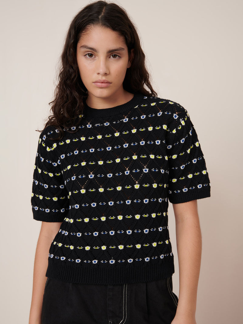 A woman with long dark hair wears the Kowtow Rhode Top – Daisy Jacquard, a black short-sleeve knit sweater with a yellow and white pattern, paired with black pants against a beige background. See our size guide for standard fit and measurements.