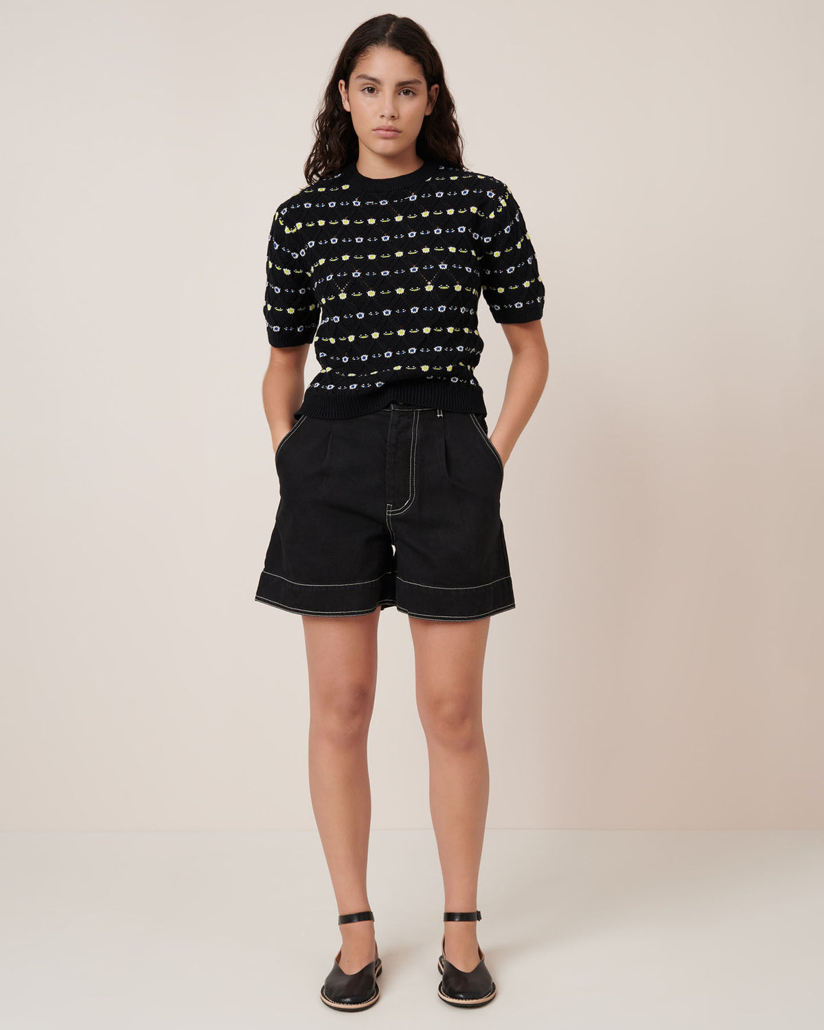 A person wears the Kowtow Rhode Top – Daisy Jacquard, styled with black shorts featuring white stitching and black sandals for a standard fit. Check our size guide for detailed garment measurements.