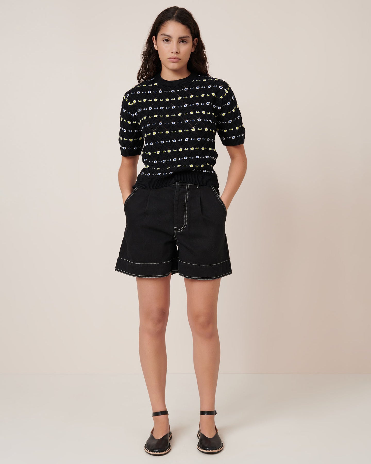 A woman with long dark hair wears the Kowtow Rhode Top – Daisy Jacquard, a black short-sleeve knit sweater with a yellow and white pattern, paired with black pants against a beige background. See our size guide for standard fit and measurements.