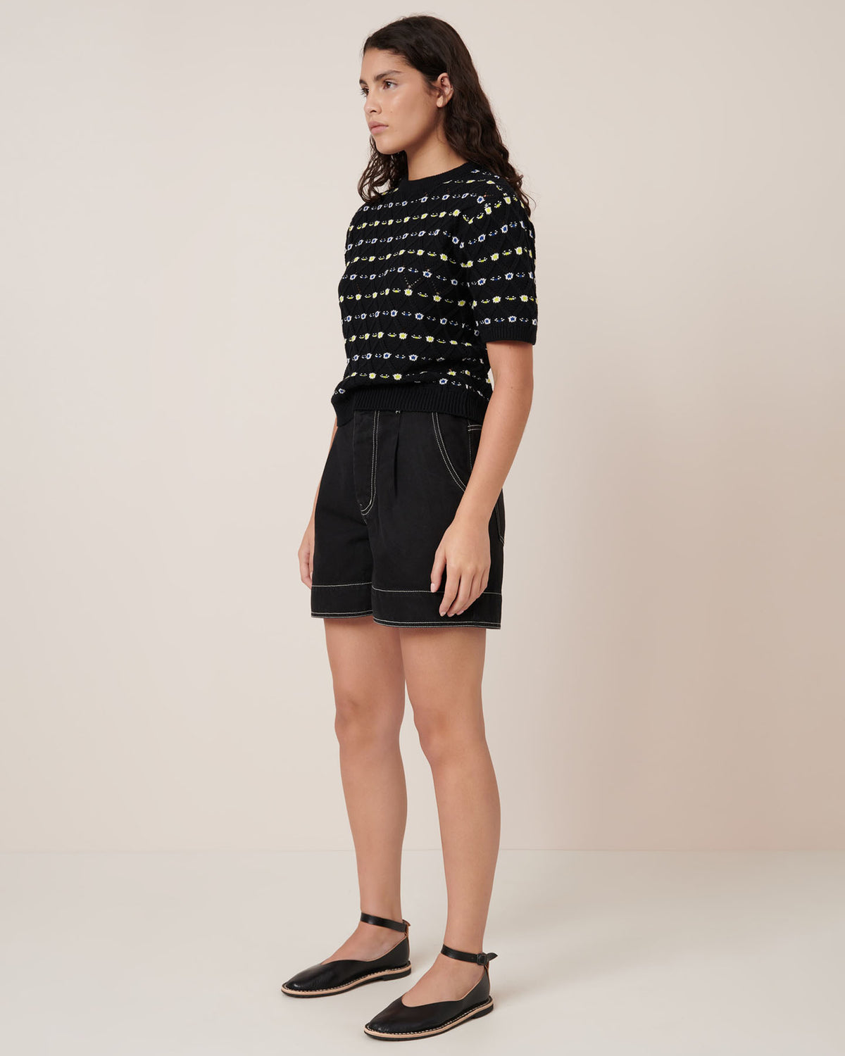 A person stands against a plain background wearing the Kowtow Rhode Top – Daisy Jacquard in a standard fit, paired with black shorts featuring white stitching and black flat shoes with ankle straps.