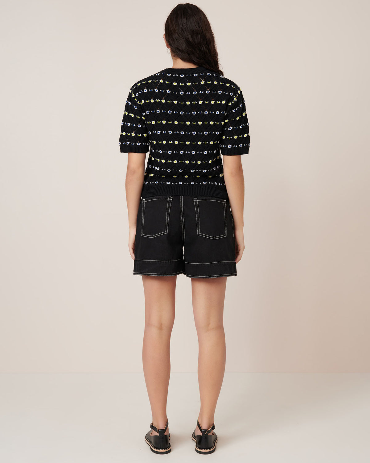 A person faces away, wearing Kowtow’s Rhode Top – Daisy Jacquard (black patterned short-sleeve sweater) and black shorts with white stitching for a standard fit. Consult our size guide for details. Black sandals finish the look against a light background.