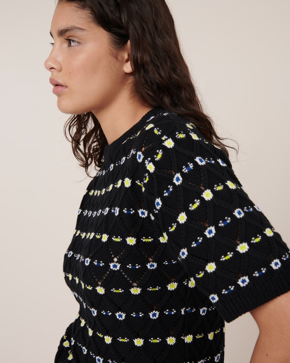 A person with long dark hair wears the Kowtow Rhode Top – Daisy Jacquard, a standard-fit black knit with blue, yellow, and white geometric patterns, standing against a plain beige background.