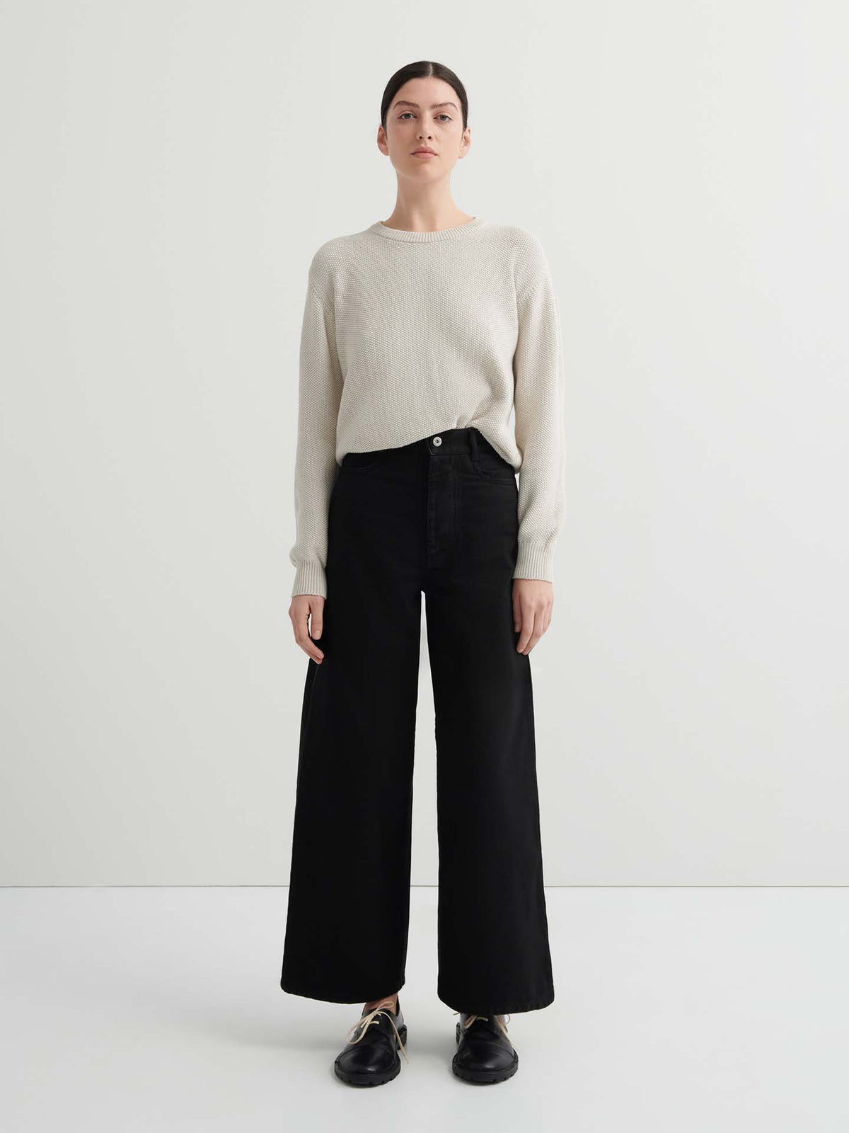 A person stands facing forward in a relaxed fit light beige sweater, Sailor Jeans – Black Denim, and black shoes against a plain white background.