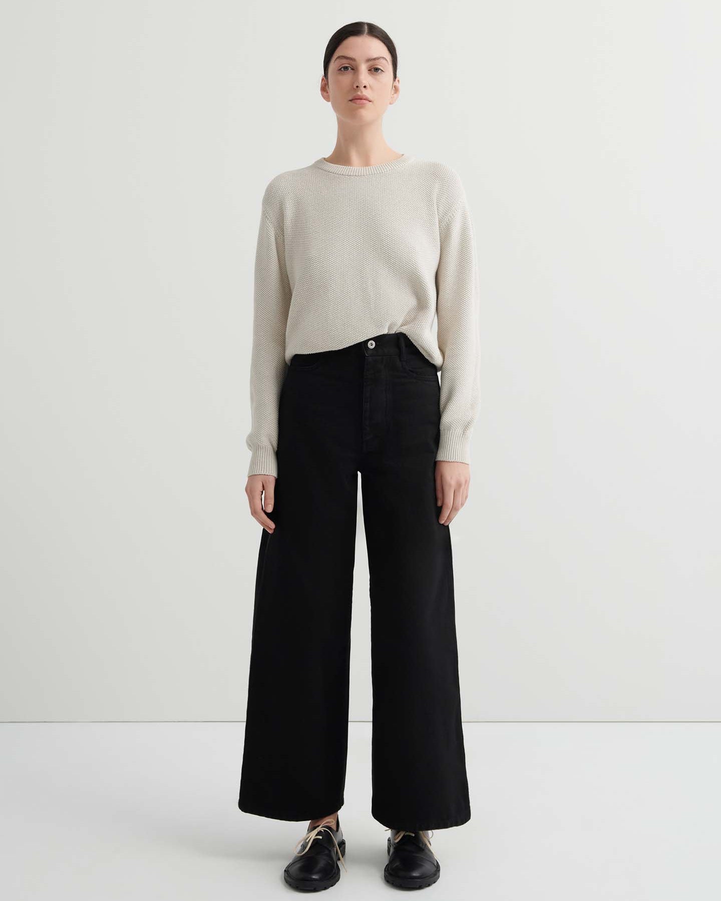 A person stands facing forward in a relaxed fit light beige sweater, Sailor Jeans – Black Denim, and black shoes against a plain white background.