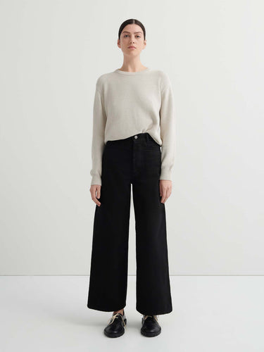 A person stands facing forward in a relaxed fit light beige sweater, Sailor Jeans – Black Denim, and black shoes against a plain white background.