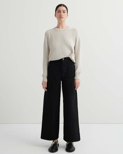 A person stands facing forward in a relaxed fit light beige sweater, Sailor Jeans – Black Denim, and black shoes against a plain white background.