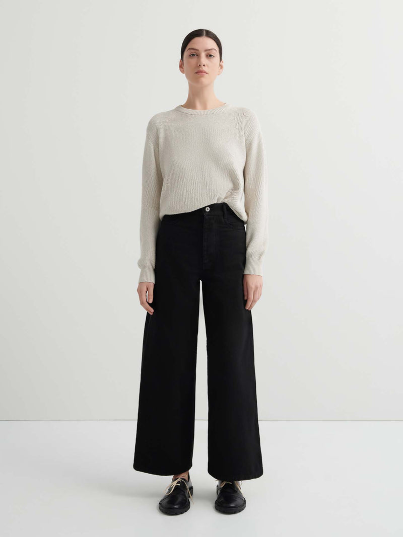 A person stands facing forward in a relaxed fit light beige sweater, Sailor Jeans – Black Denim, and black shoes against a plain white background.