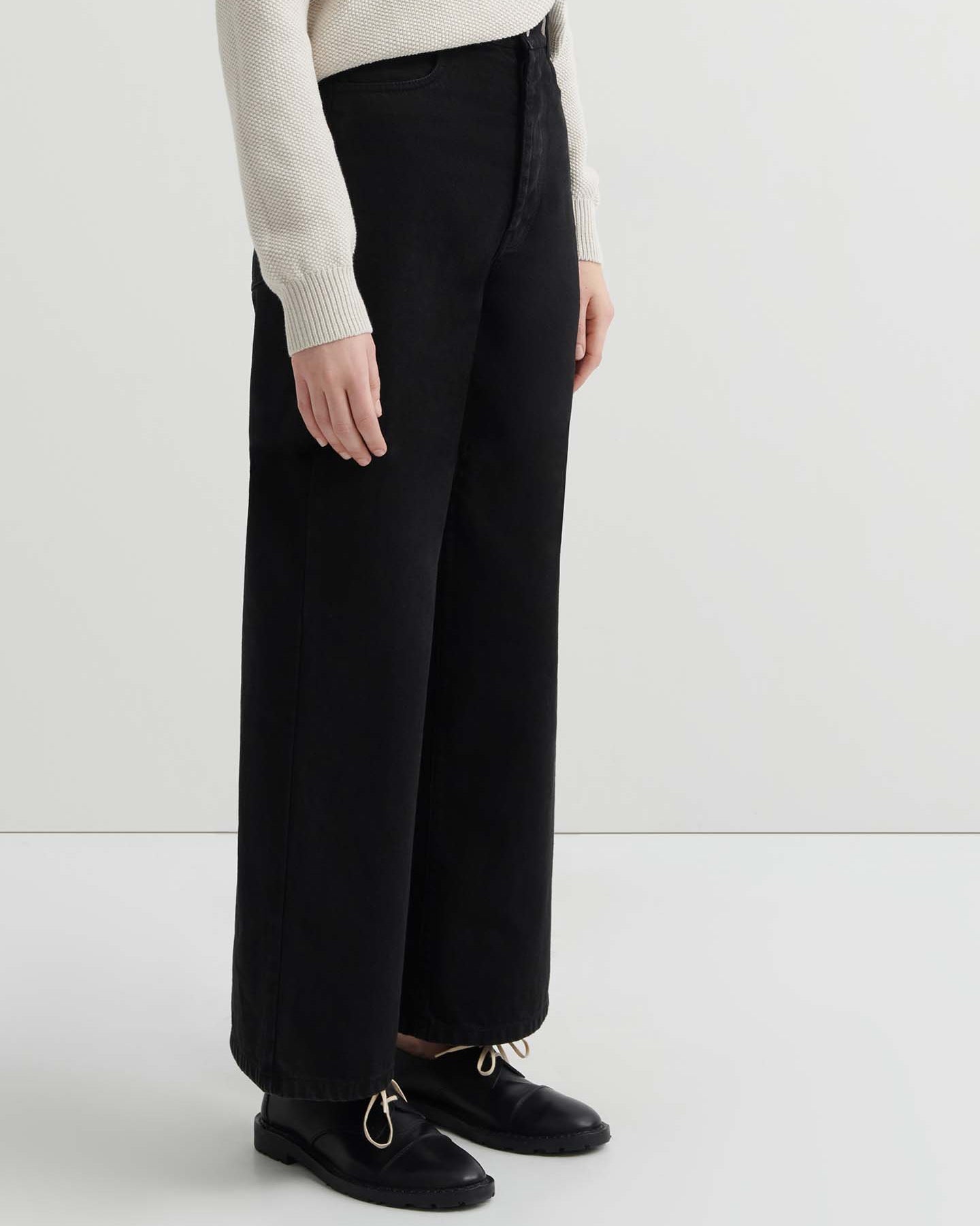 A person stands facing forward in a relaxed fit light beige sweater, Sailor Jeans – Black Denim, and black shoes against a plain white background.