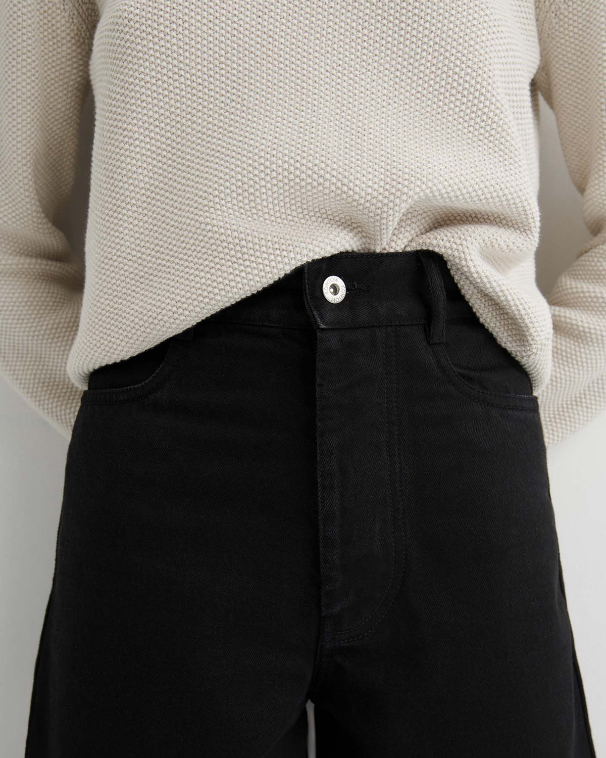 Person wearing textured off-white sweater and Sailor Jeans – Black Denim in a relaxed fit, standing with hands behind their back.