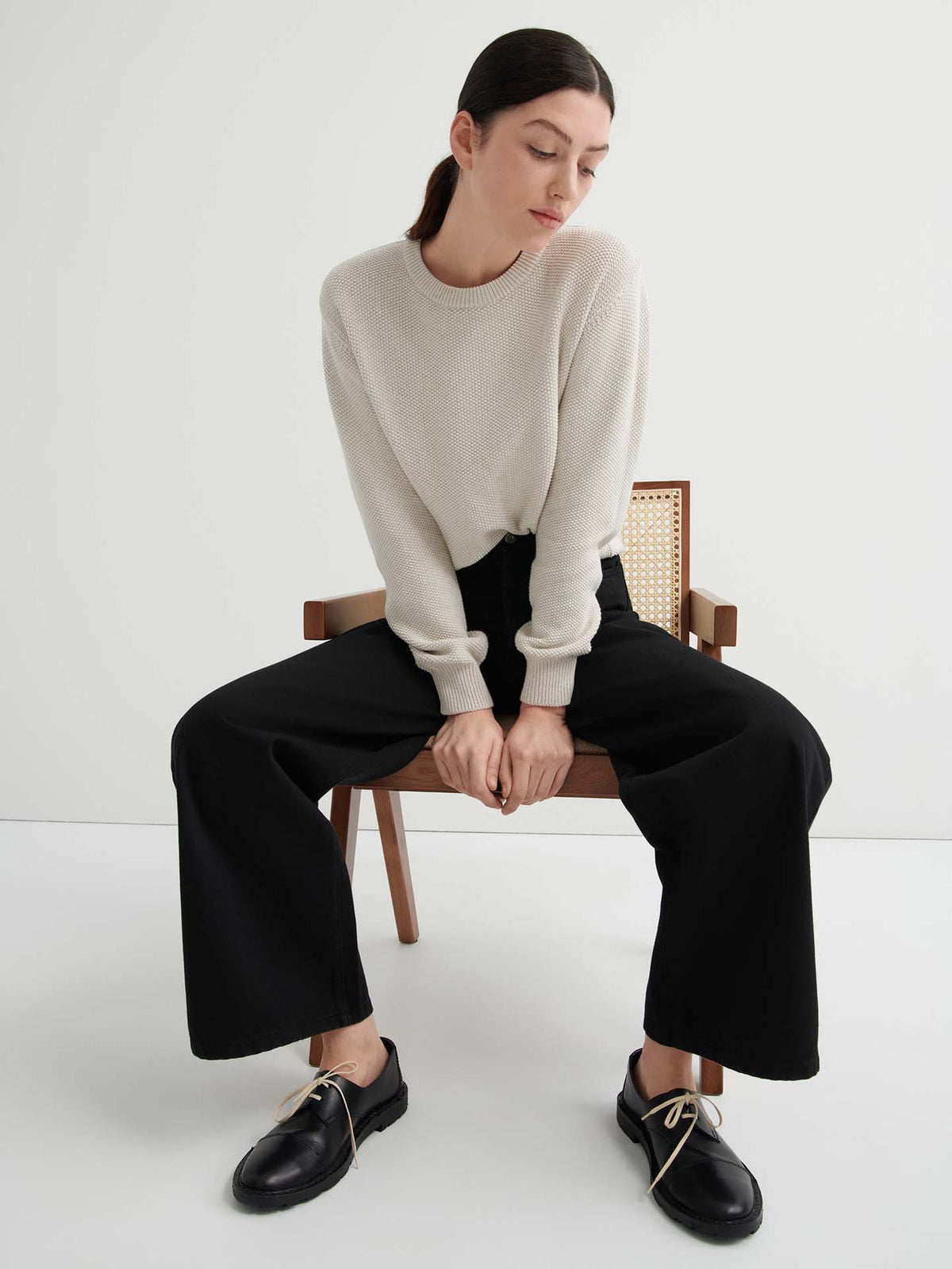A woman sits on a wooden chair wearing a cream sweater, Sailor Jeans – Black Denim, and black shoes with beige laces against a plain white background.