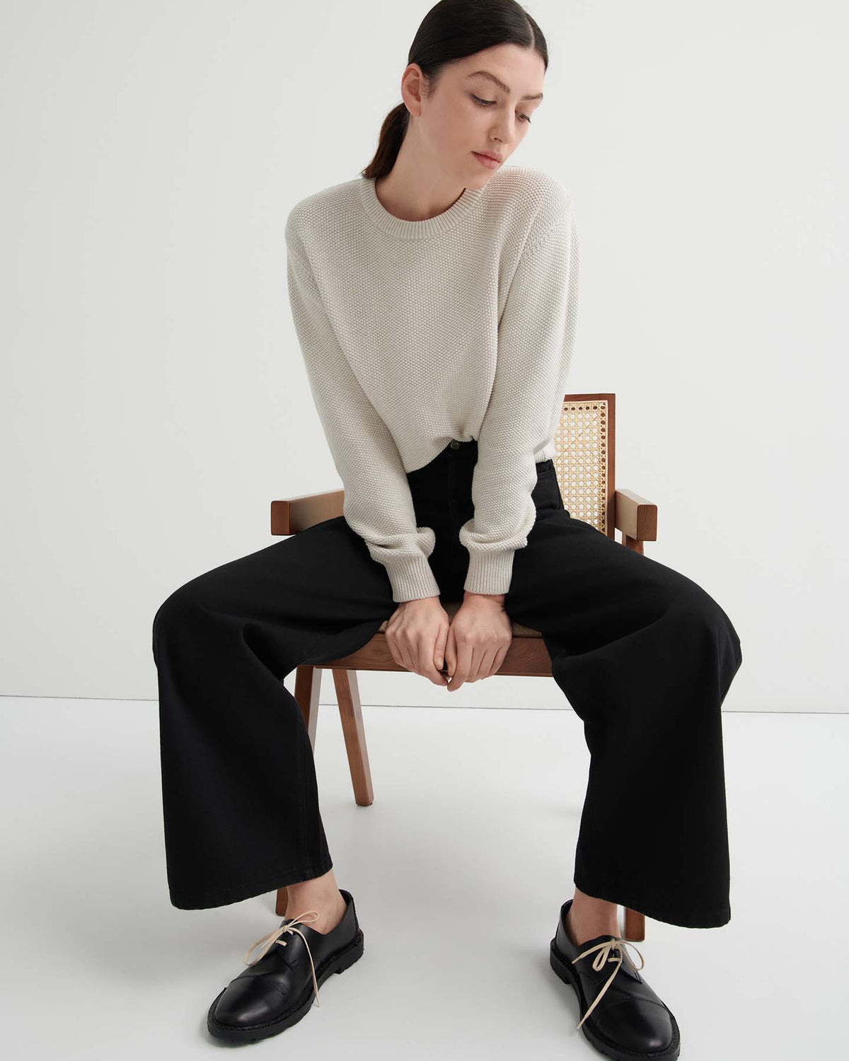 A woman sits on a wooden chair wearing a cream sweater, Sailor Jeans – Black Denim, and black shoes with beige laces against a plain white background.