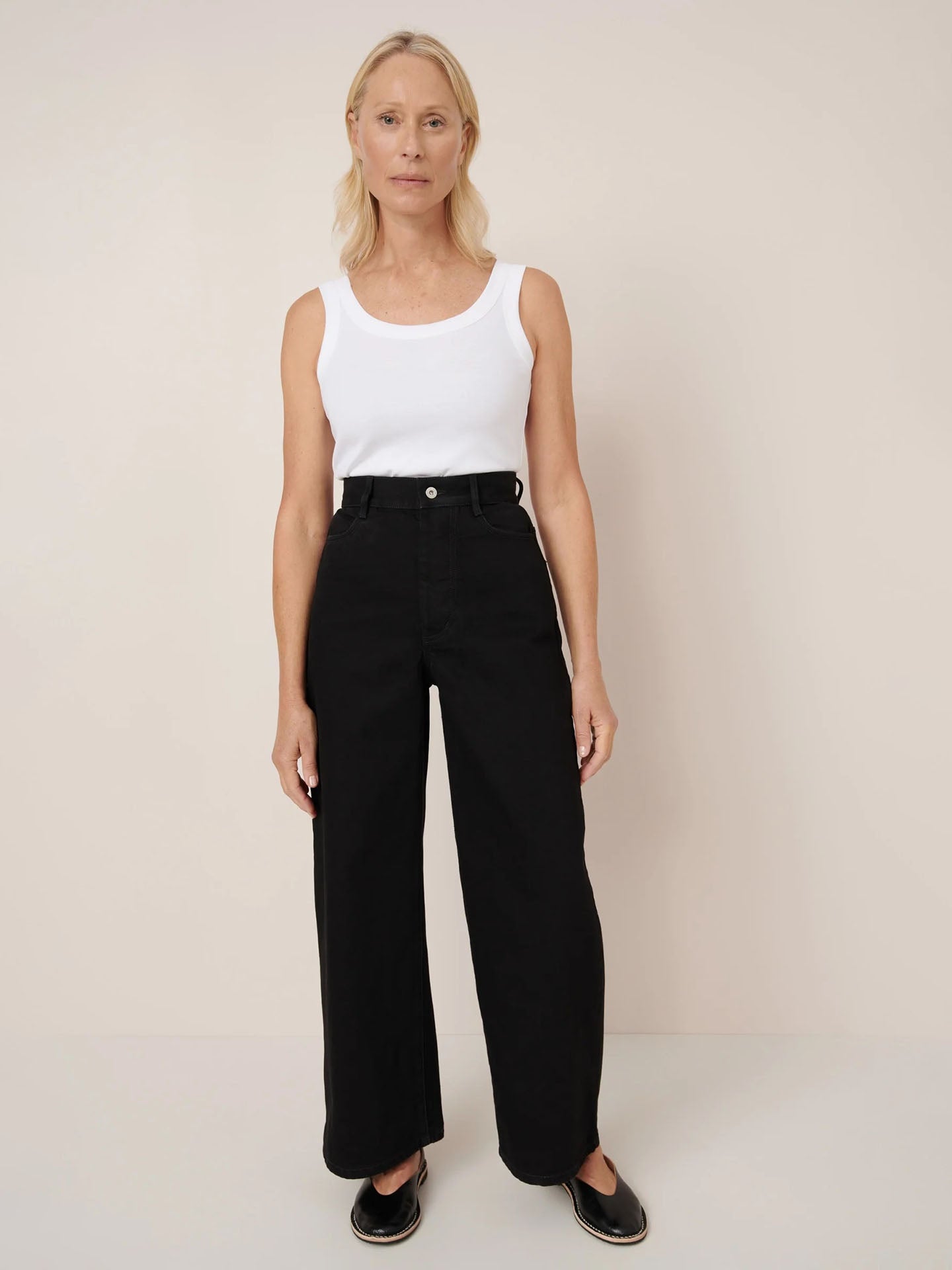 A woman stands against a plain background wearing a white sleeveless top, Kowtow Sailor Jeans in black denim, and black shoes.