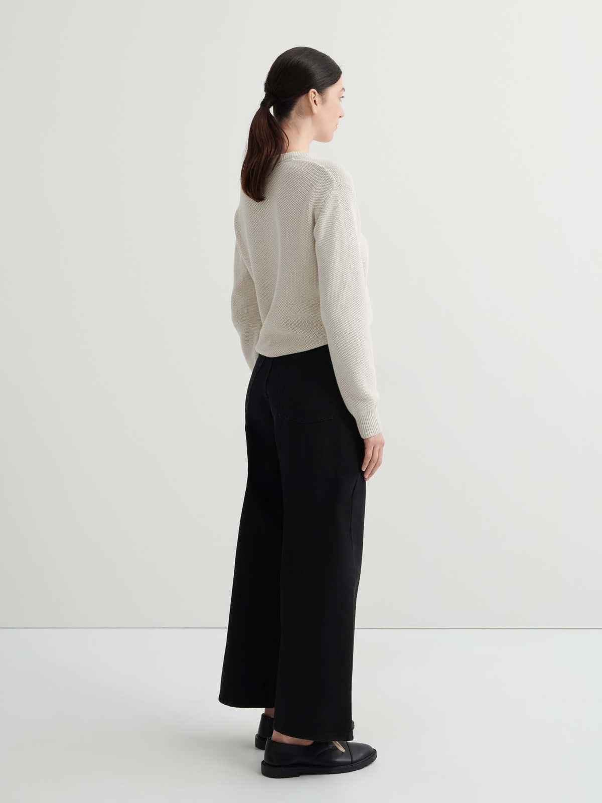 A woman faces away in a light beige sweater, Sailor Jeans – Black Denim with a relaxed wide-leg fit, and black shoes against a plain white background.
