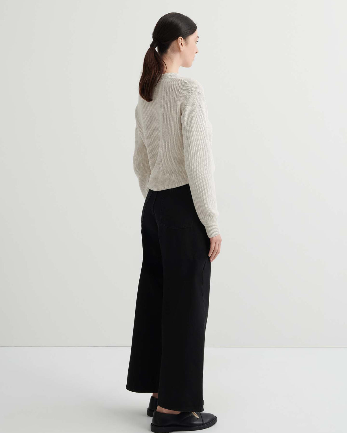 A woman faces away in a light beige sweater, Sailor Jeans – Black Denim with a relaxed wide-leg fit, and black shoes against a plain white background.