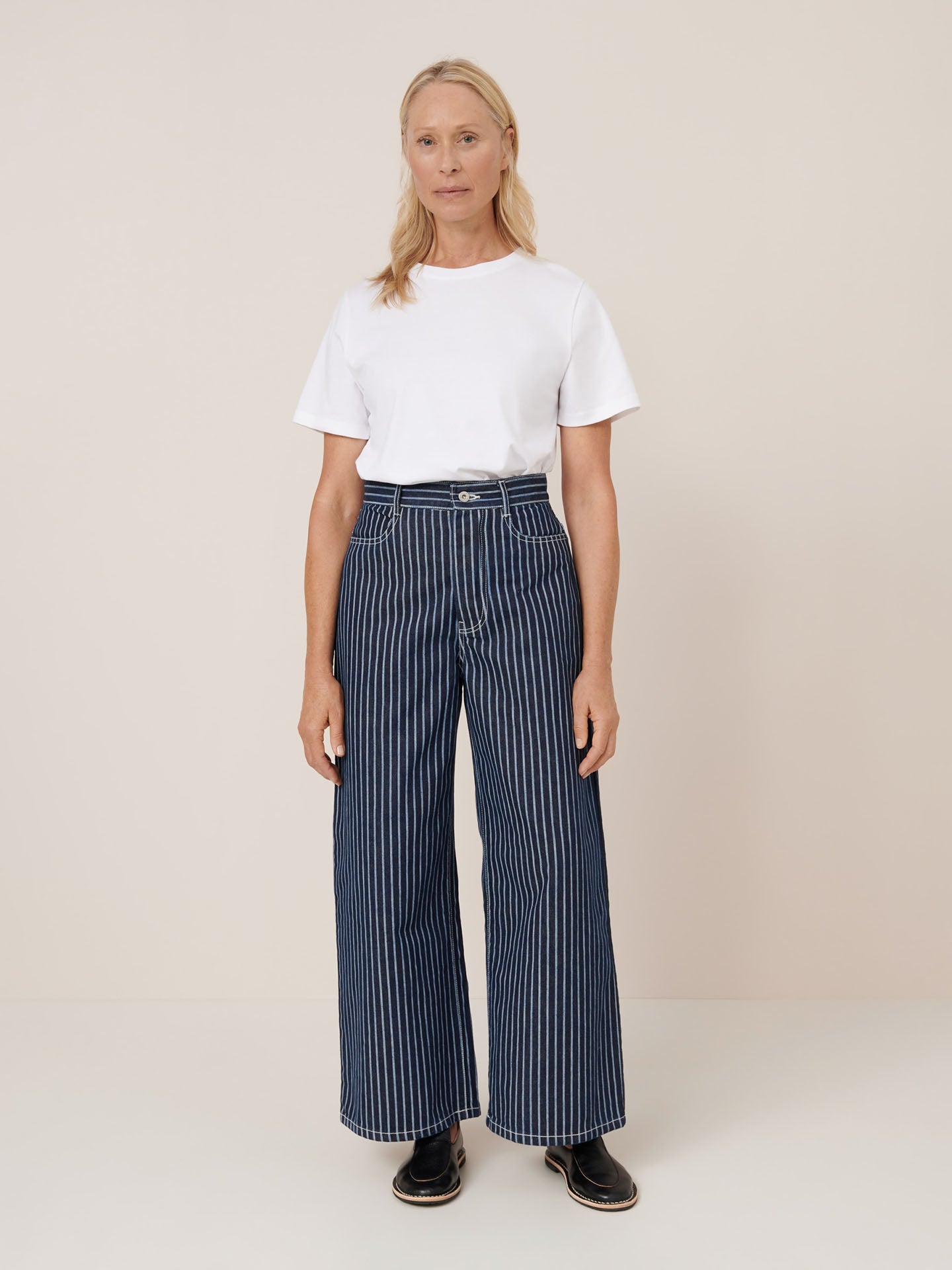 A woman stands against a plain background wearing a white t-shirt, black shoes, and Kowtow Sailor Jeans – Chalk Stripe in a relaxed high-waist fit.