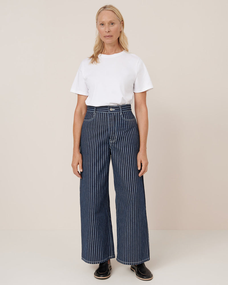 A woman stands against a plain background wearing a white t-shirt, black shoes, and Kowtow Sailor Jeans – Chalk Stripe in a relaxed high-waist fit.