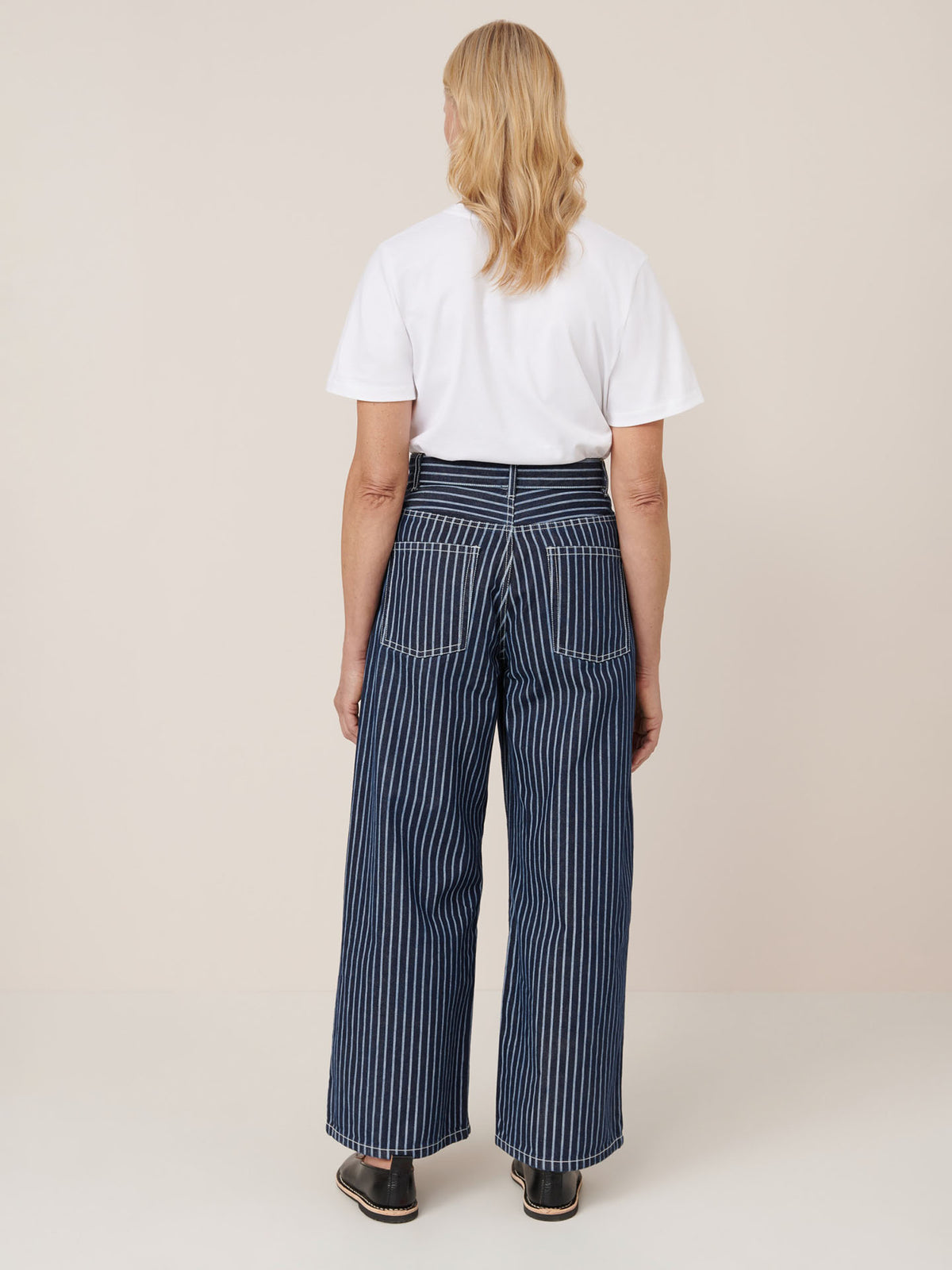 A person faces away, wearing a white t-shirt and Kowtow Sailor Jeans – Chalk Stripe: high-waisted, wide-leg, blue and white striped jeans made from pre-washed fabric, paired with black shoes against a plain background.