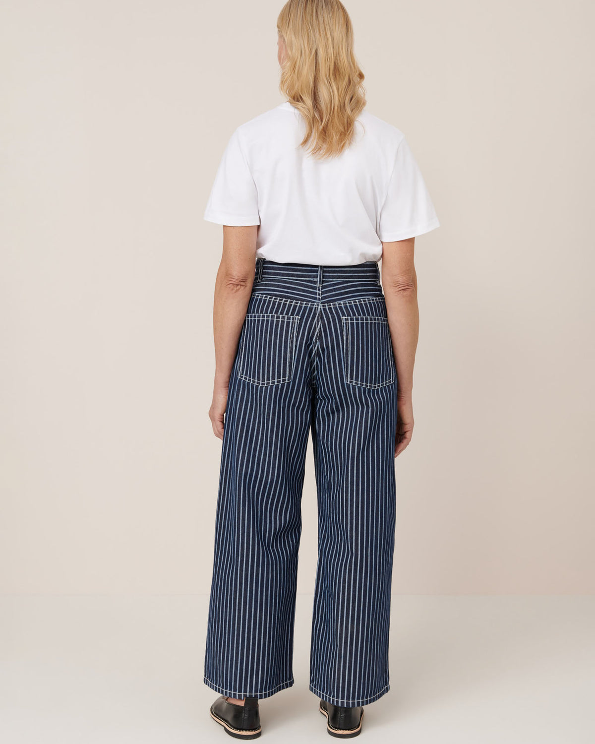 A person faces away, wearing a white t-shirt and Kowtow Sailor Jeans – Chalk Stripe: high-waisted, wide-leg, blue and white striped jeans made from pre-washed fabric, paired with black shoes against a plain background.