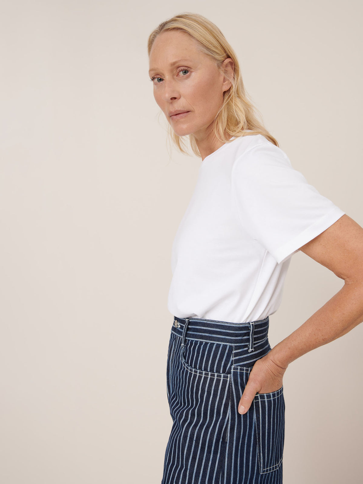 A woman stands sideways with one hand in her pocket, wearing a white t-shirt and Kowtow Sailor Jeans – Chalk Stripe, featuring high waist and relaxed fit denim with blue and white stripes against a plain background.