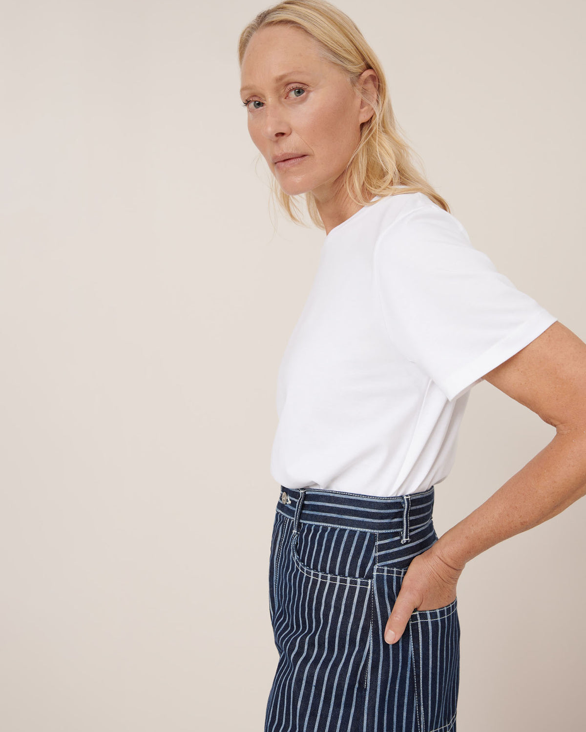A woman stands sideways with one hand in her pocket, wearing a white t-shirt and Kowtow Sailor Jeans – Chalk Stripe, featuring high waist and relaxed fit denim with blue and white stripes against a plain background.