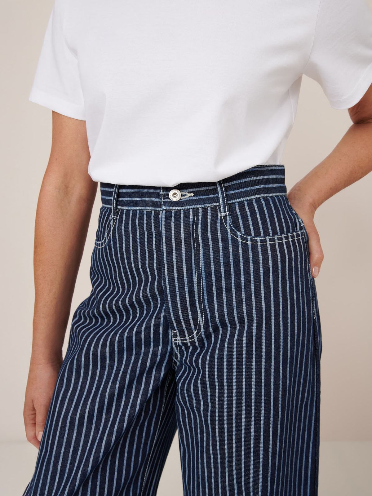 A person wears Kowtow Sailor Jeans – Chalk Stripe, high-waisted navy blue jeans with white pinstripes, paired with a white short-sleeve shirt, standing with one hand on their hip.