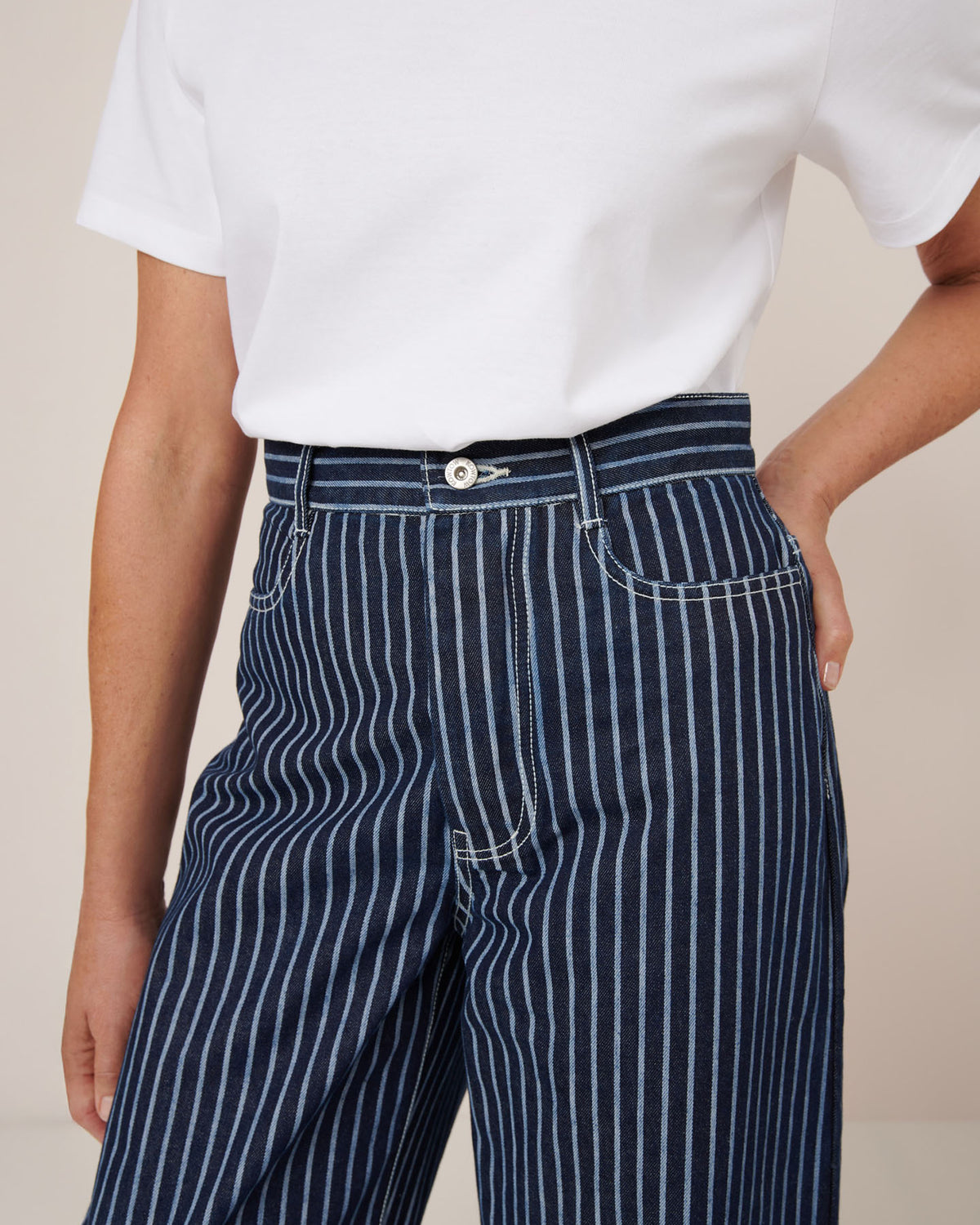 A person wears Kowtow Sailor Jeans – Chalk Stripe, high-waisted navy blue jeans with white pinstripes, paired with a white short-sleeve shirt, standing with one hand on their hip.