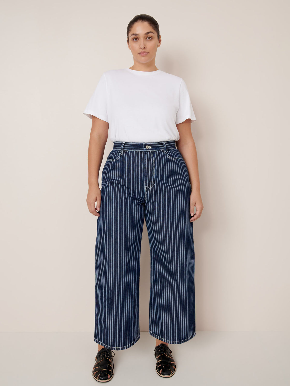 A person stands against a plain background wearing a white t-shirt, Kowtow Sailor Jeans – Chalk Stripe with a relaxed fit, and black sandals.