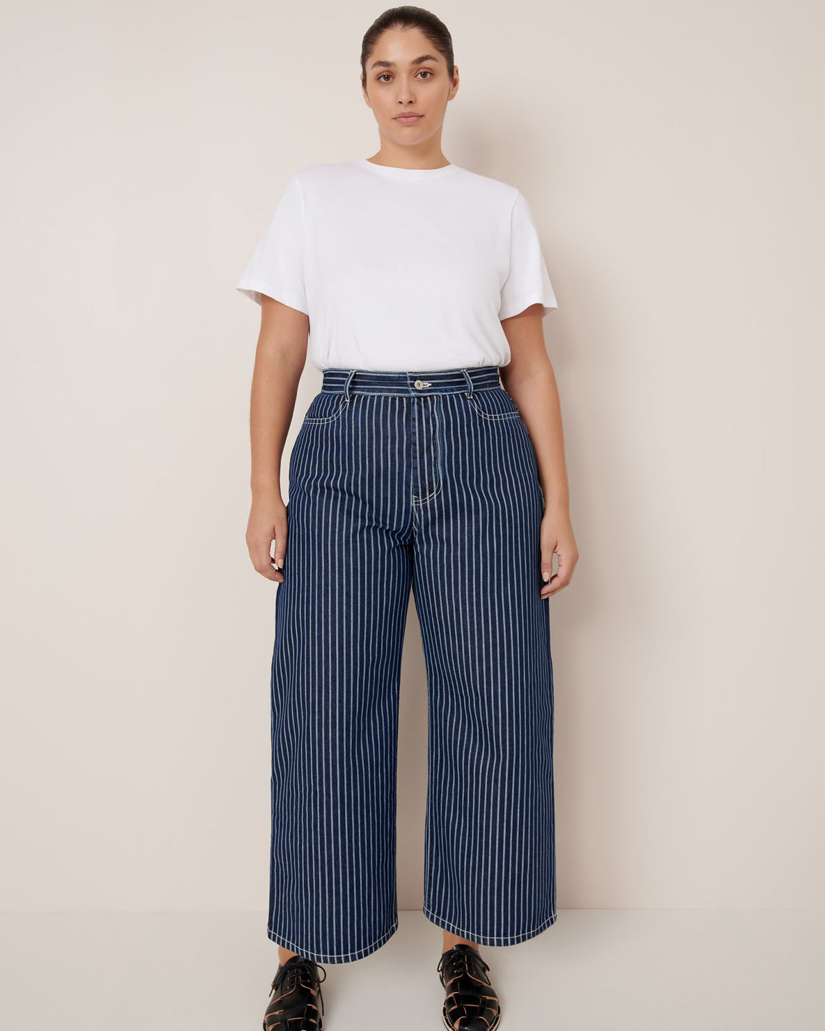 A person stands against a plain background wearing a white t-shirt, Kowtow Sailor Jeans – Chalk Stripe with a relaxed fit, and black sandals.