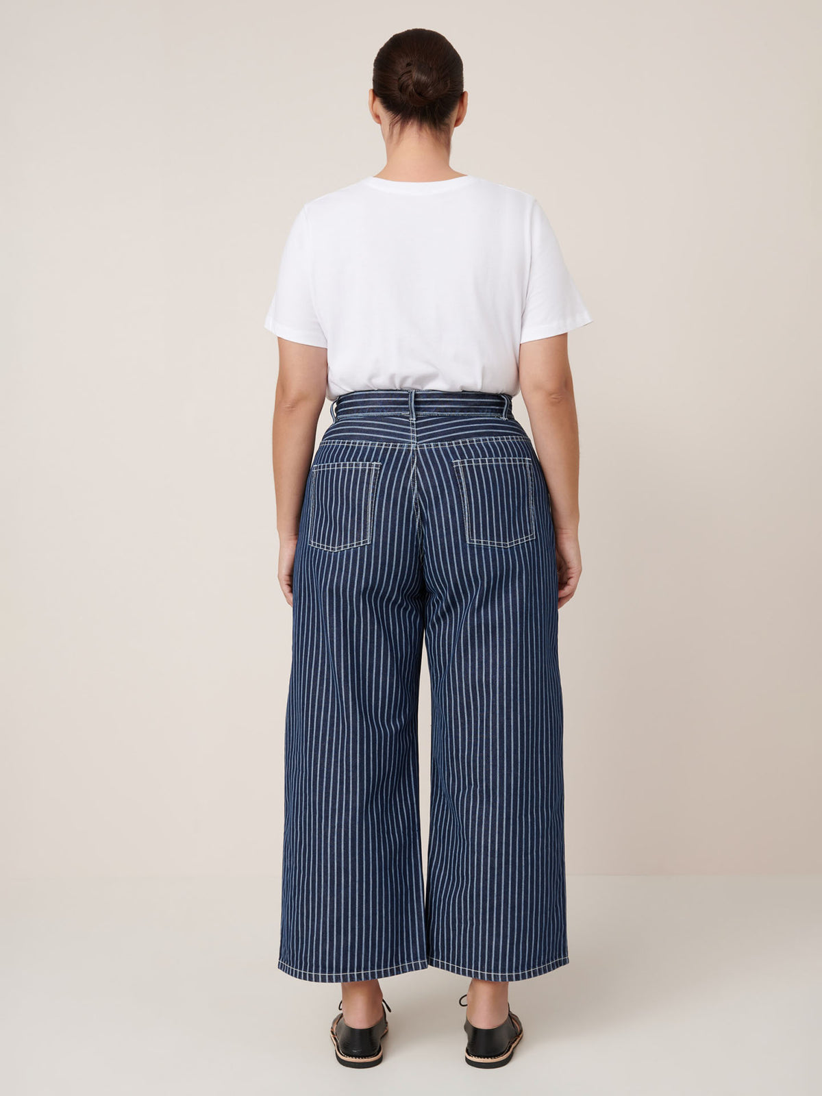 A person stands with their back to the camera, wearing a white T-shirt and Kowtow Sailor Jeans – Chalk Stripe, styled with black sandals against a plain beige background.