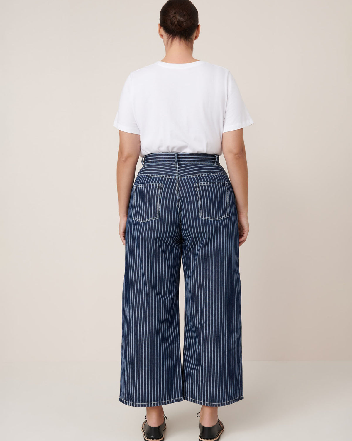A person stands with their back to the camera, wearing a white T-shirt and Kowtow Sailor Jeans – Chalk Stripe, styled with black sandals against a plain beige background.