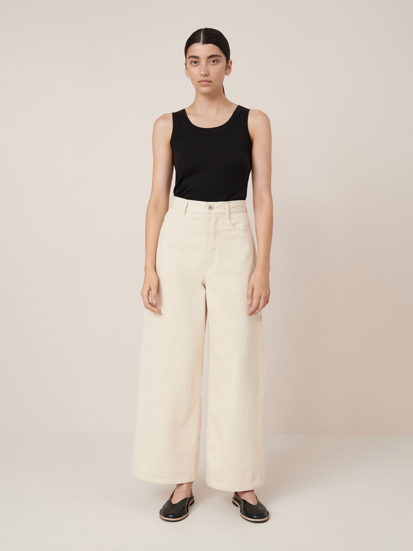 A person stands against a plain background wearing a black sleeveless top, Kowtow Sailor Jeans in ecru denim with a relaxed wide-leg fit, and black slip-on shoes.