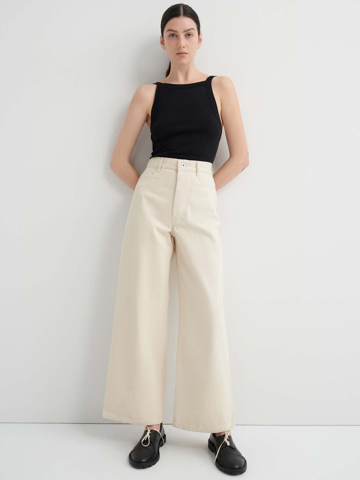 A woman stands against a white wall wearing a black sleeveless top, Sailor Jeans in ecru denim with a high waist and relaxed fit, and black shoes with gold details.