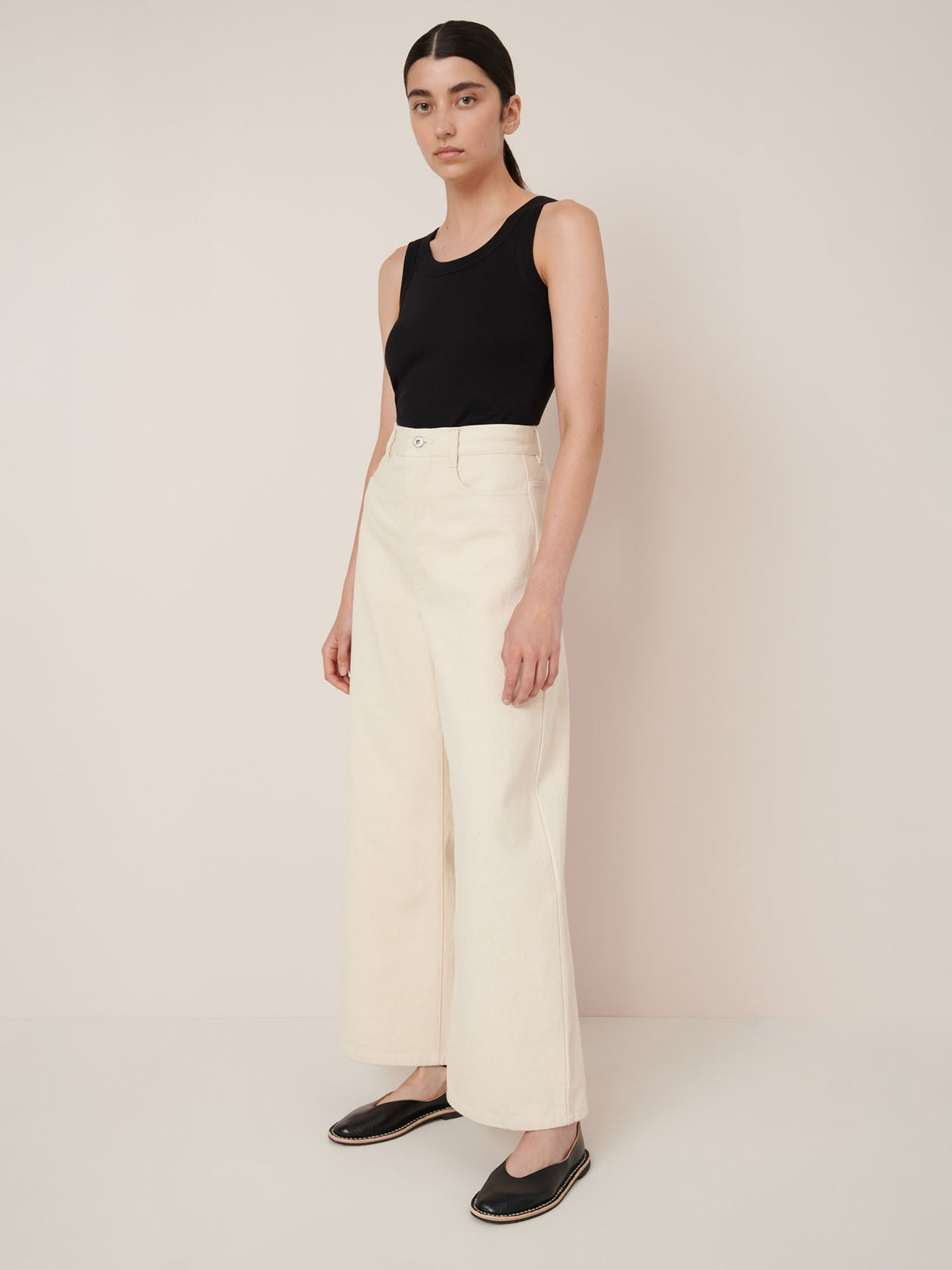 A woman stands against a plain wall wearing a black sleeveless top, Kowtow Sailor Jeans in ecru denim, and black flat shoes.