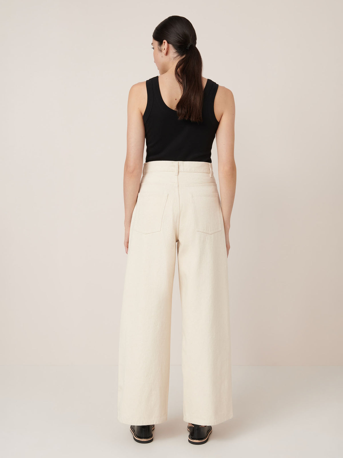 A woman faces away, wearing Kowtow&#39;s Sailor Jeans in ecru denim, paired with a black sleeveless top and black shoes against a plain light background.